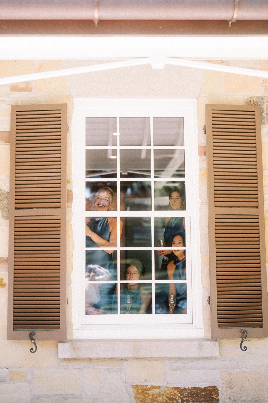Bridesmaids smiling behind a white framed window with brown shutters during a private estate wedding near Chatham-Kent, Ontario.