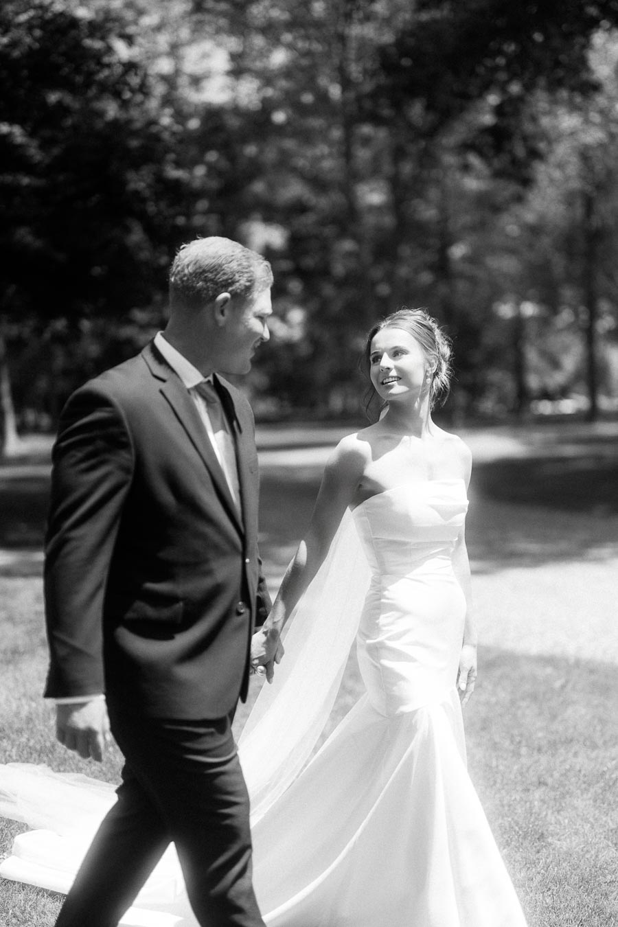 Black and white wedding portrait of a bride and groom holding hands while walking outdoors on a sunny lawn with trees.