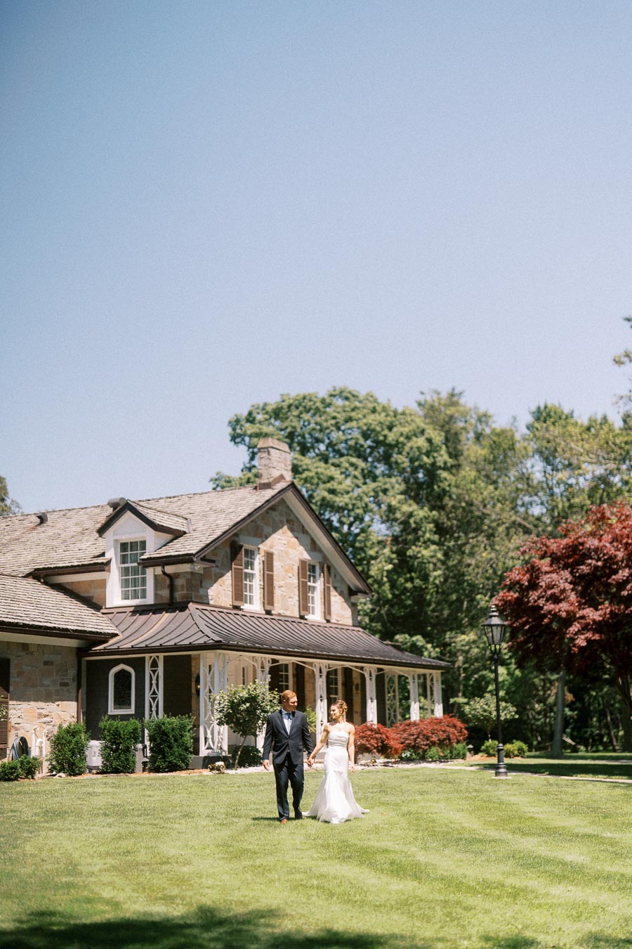 Bride and groom holding hands on the lawn of a private estate near Chatham-Kent, Ontario, with a stone house and trees behind them.