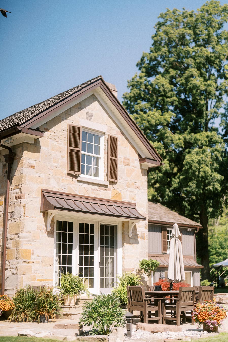 Stone house patio with outdoor dining table, closed umbrella, flowering planters, and trees on a sunny private estate in Chatham-Kent, Ontario.