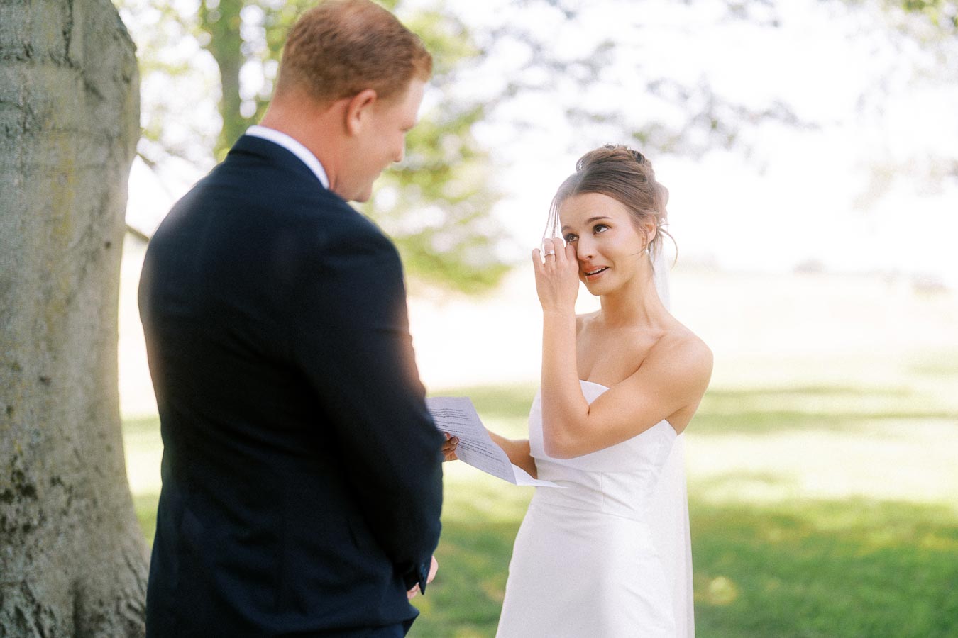Bride wipes a tear while reading vows with groom under a tree at an outdoor private estate wedding near Chatham-Kent, Ontario.