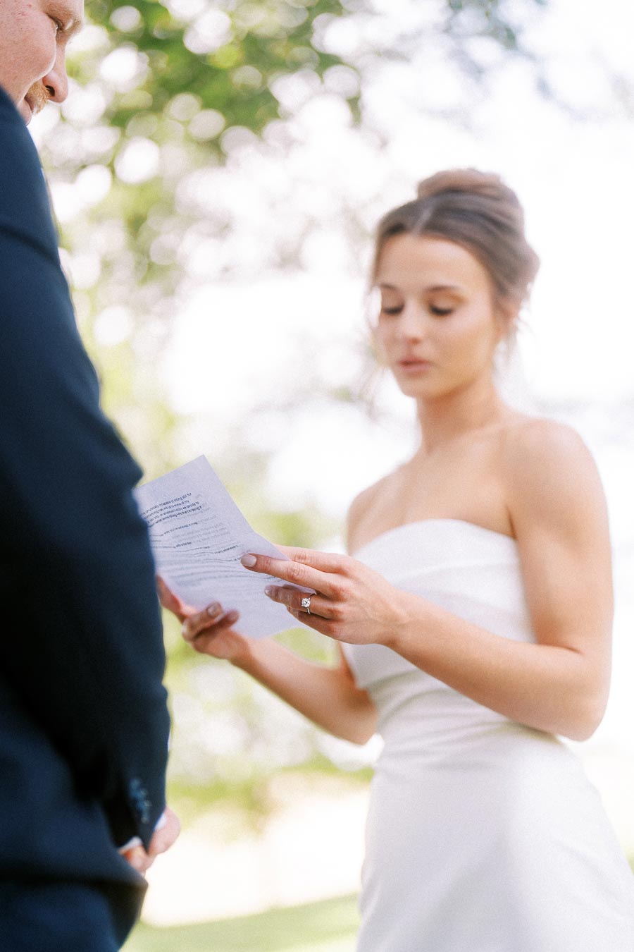 Bride in a white strapless dress reads wedding vows from paper to groom under trees during an outdoor private estate wedding near Chatham-Kent, Ontario.