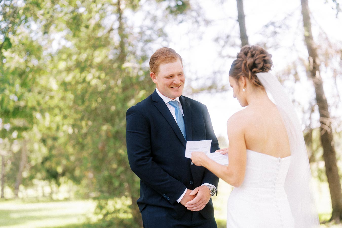 Bride reads wedding vows to smiling groom beneath trees during an outdoor private estate wedding in Chatham-Kent, Ontario, Canada.