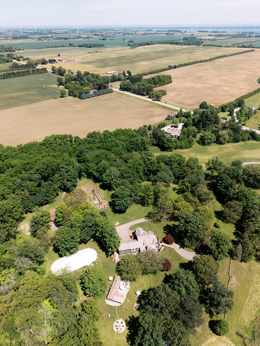 Aerial view of a private estate wedding tent and farmhouse surrounded by trees, lawns, farmland, and wind turbines near Chatham-Kent, Ontario, Canada.