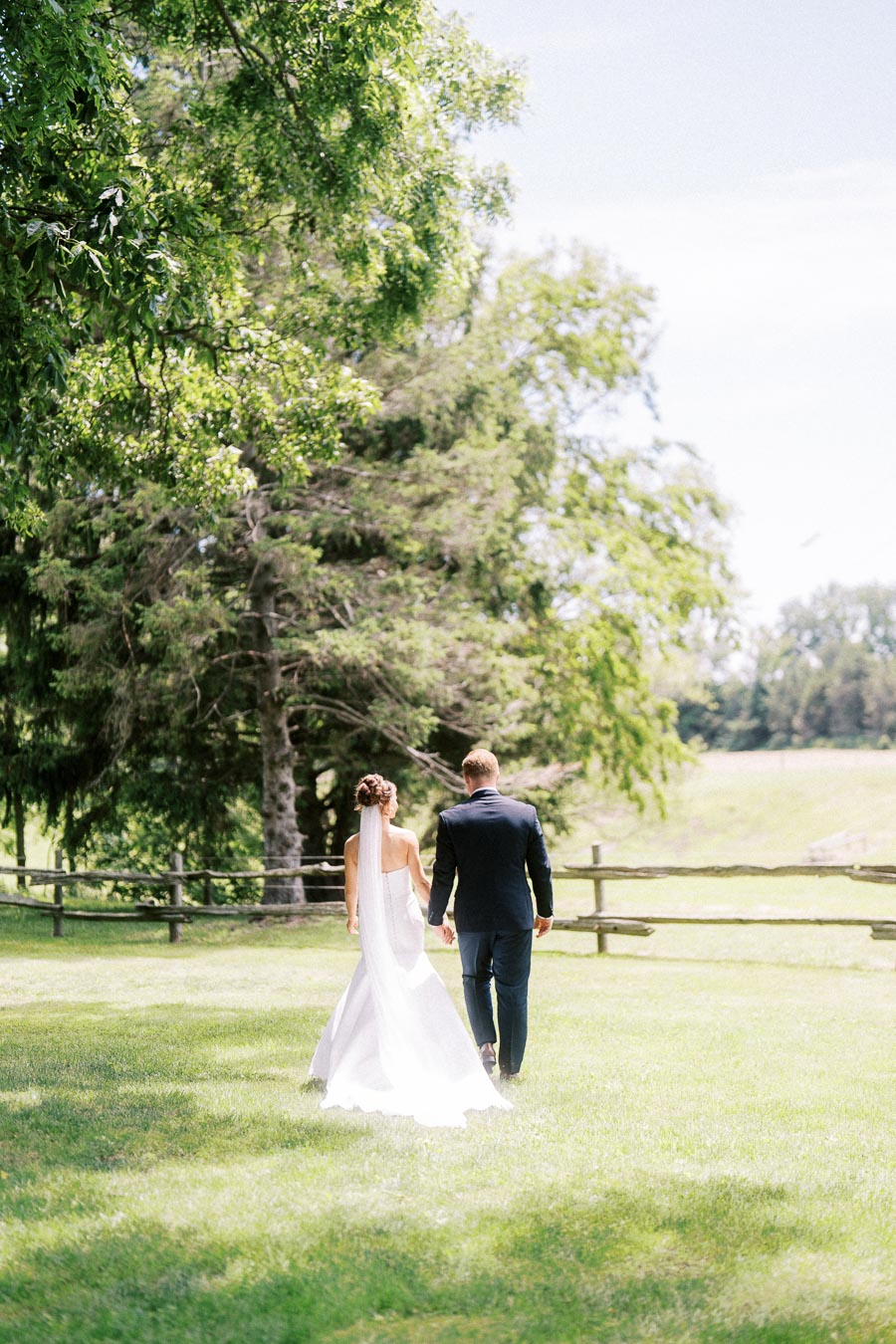 Bride and groom walking hand in hand across a grassy private estate lawn under trees near Chatham-Kent, Ontario.