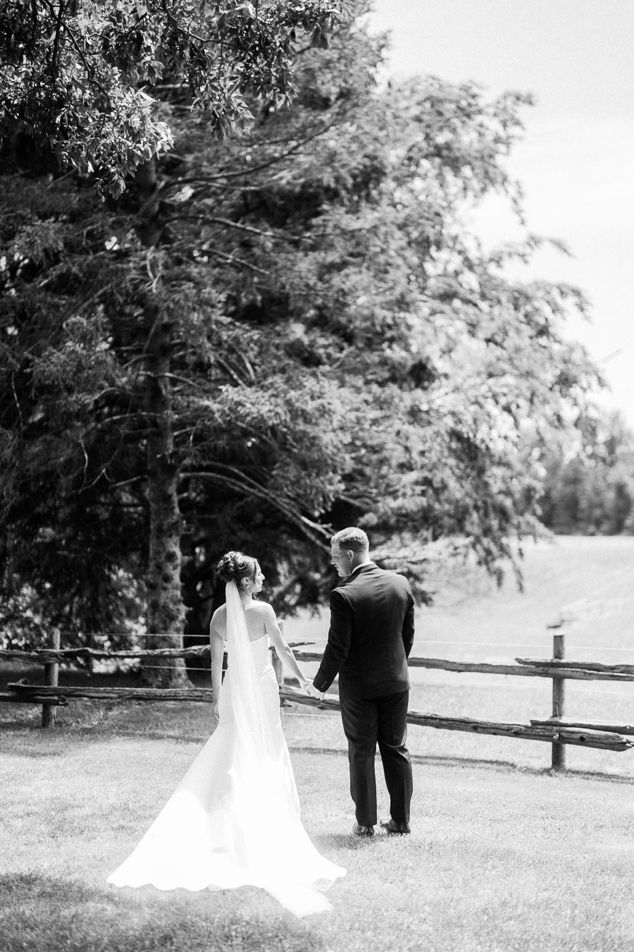 Black and white wedding portrait of a bride and groom holding hands near a wooden fence on a farm estate in Chatham-Kent, Ontario.