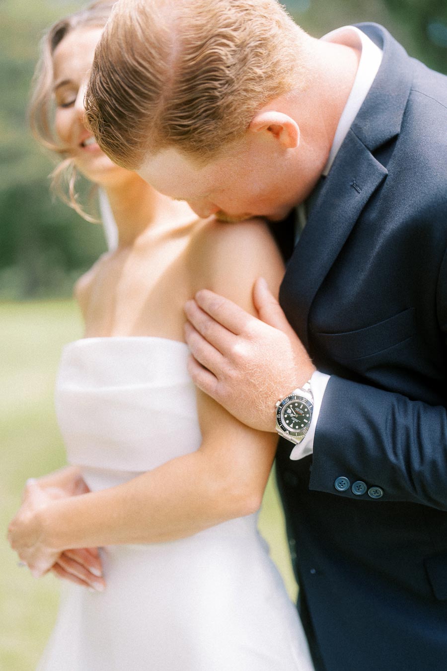 Groom in a dark suit kisses the bride's shoulder during an outdoor wedding portrait on a private farm estate near Chatham-Kent, Ontario.