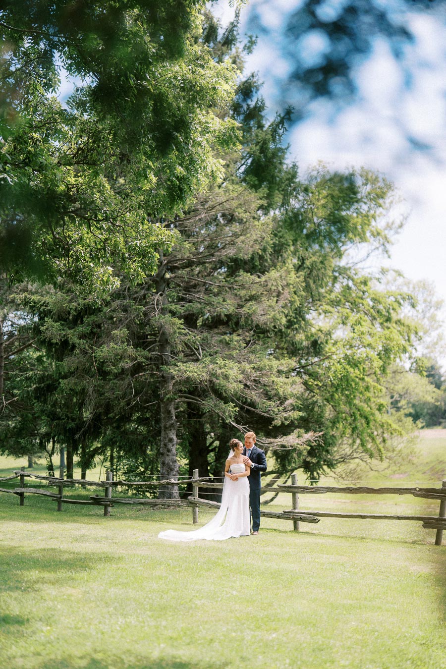 Bride and groom standing on a grassy private farm estate beside a rustic fence and tall trees near Chatham-Kent, Ontario, Canada.