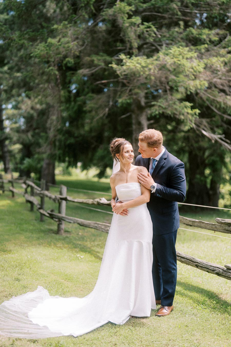 Bride and groom posing together outdoors by a rustic wooden fence on a green farm property in Ontario.
