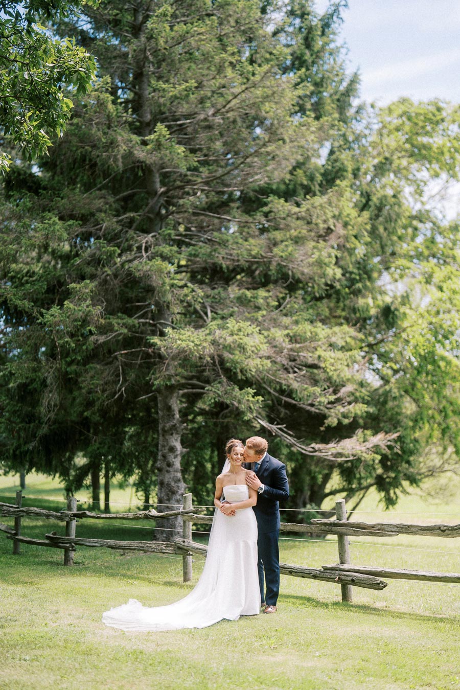Bride and groom posing together on a grassy private farm property near a rustic wooden fence and tall trees in Ontario, Canada.