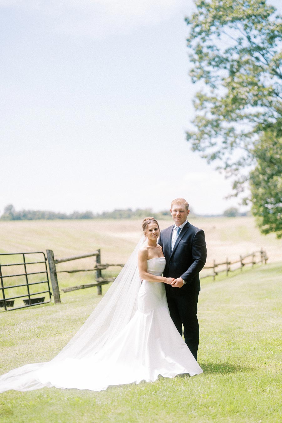 Bride and groom standing together on a grassy farm lawn near Chatham-Kent, Ontario, with fields, fence, gate, and trees in the background.