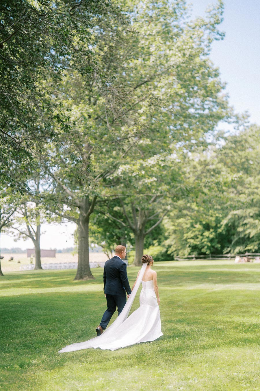 Bride and groom walking across a green lawn under large trees at a private farm wedding in Chatham-Kent, Ontario, Canada.