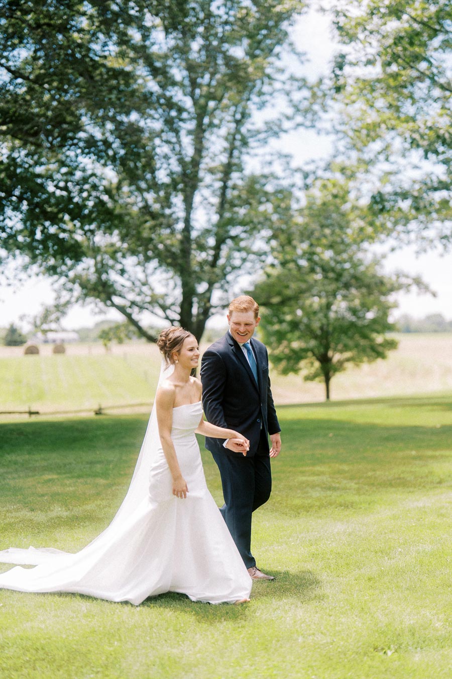 Bride and groom walking together on a grassy private farm property near Chatham-Kent, Ontario, with trees and fields in the background.