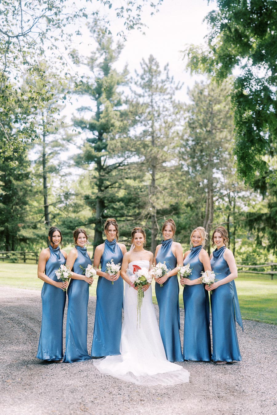 Bride with bridesmaids in blue satin dresses holding white bouquets on a private estate driveway near Chatham-Kent, Ontario.