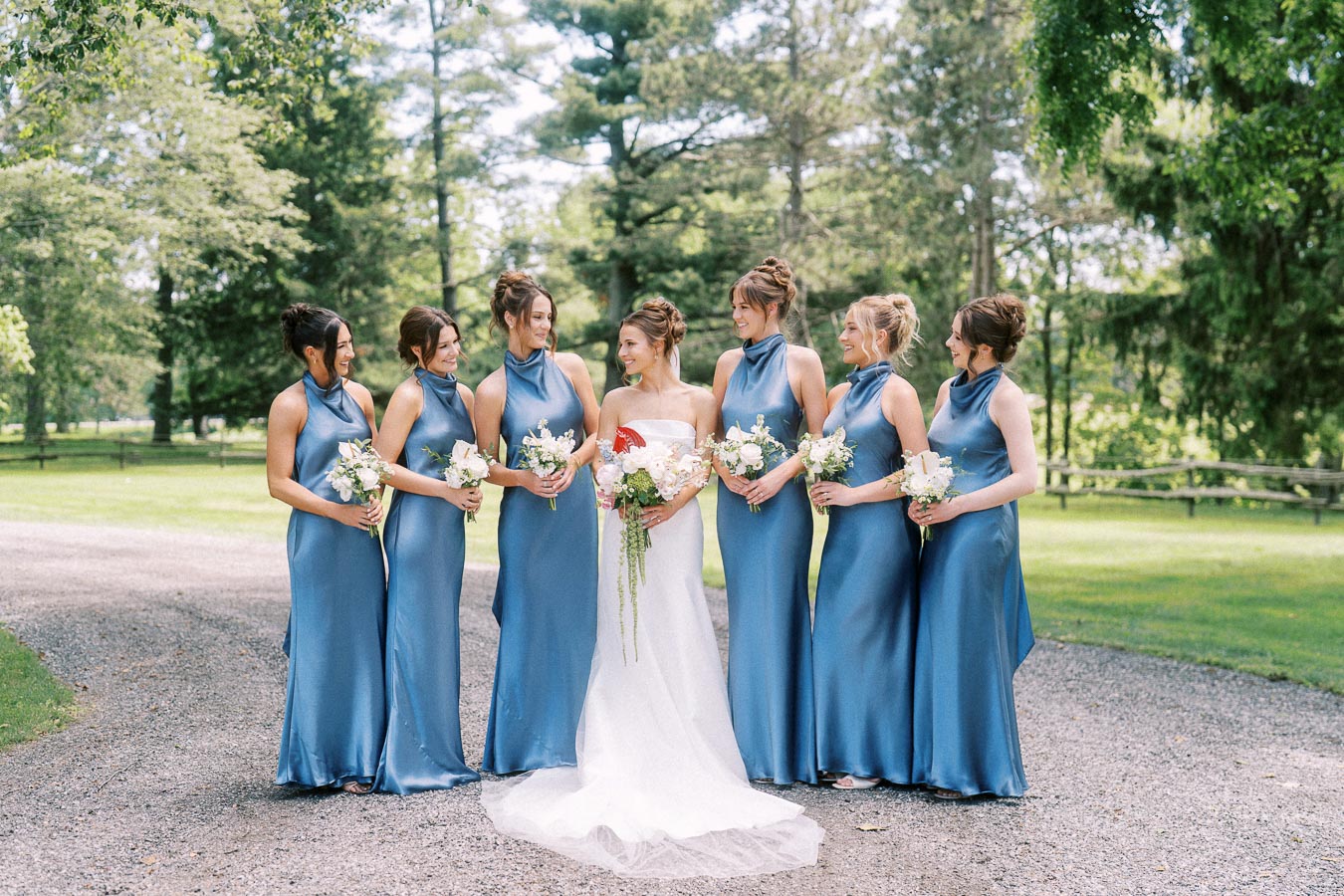 Bride with six bridesmaids in blue dresses holding bouquets on a tree lined gravel driveway in Chatham-Kent, Ontario.