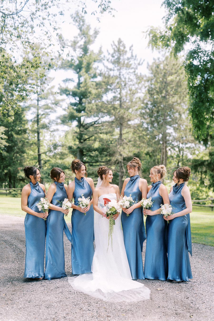 Bride with bridesmaids in blue dresses holding bouquets on a tree lined private estate near Chatham Kent, Ontario, Canada.