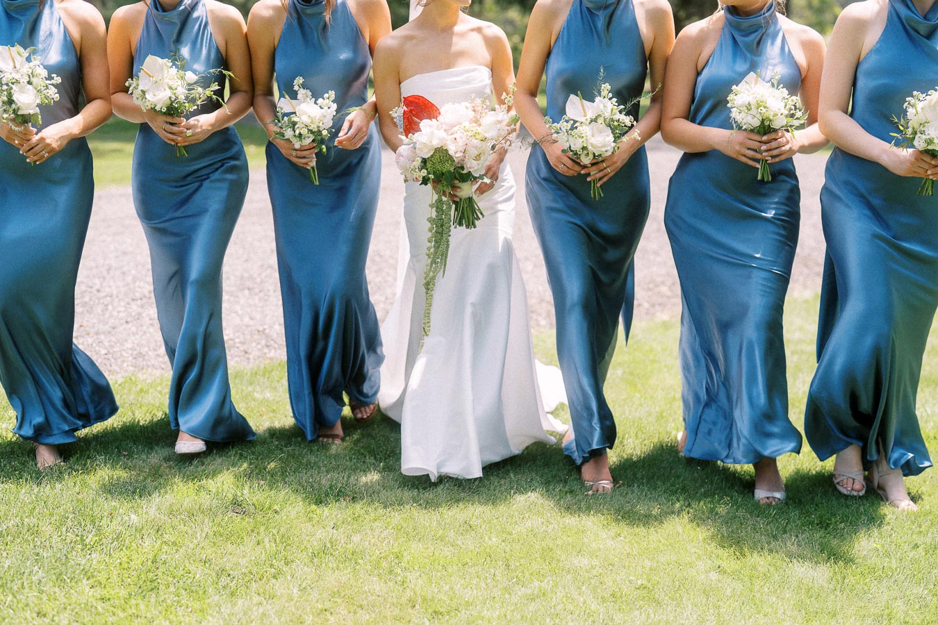 Bride and bridesmaids in blue satin dresses holding white bouquets outdoors on a lawn near Chatham-Kent, Ontario.