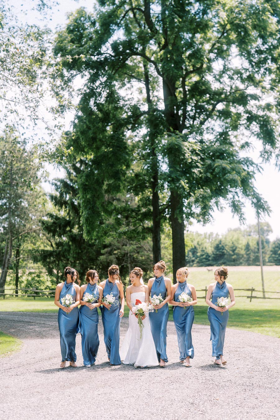 Bride walking with bridesmaids in blue dresses on a private estate driveway near Chatham-Kent, Ontario, during an outdoor wedding photo session.