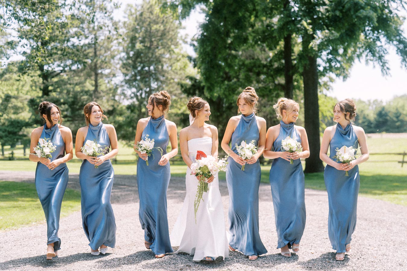 Bride with bridesmaids in blue dresses walking outdoors on a private estate near Chatham-Kent, Ontario, holding bouquets on a sunny gravel lane.