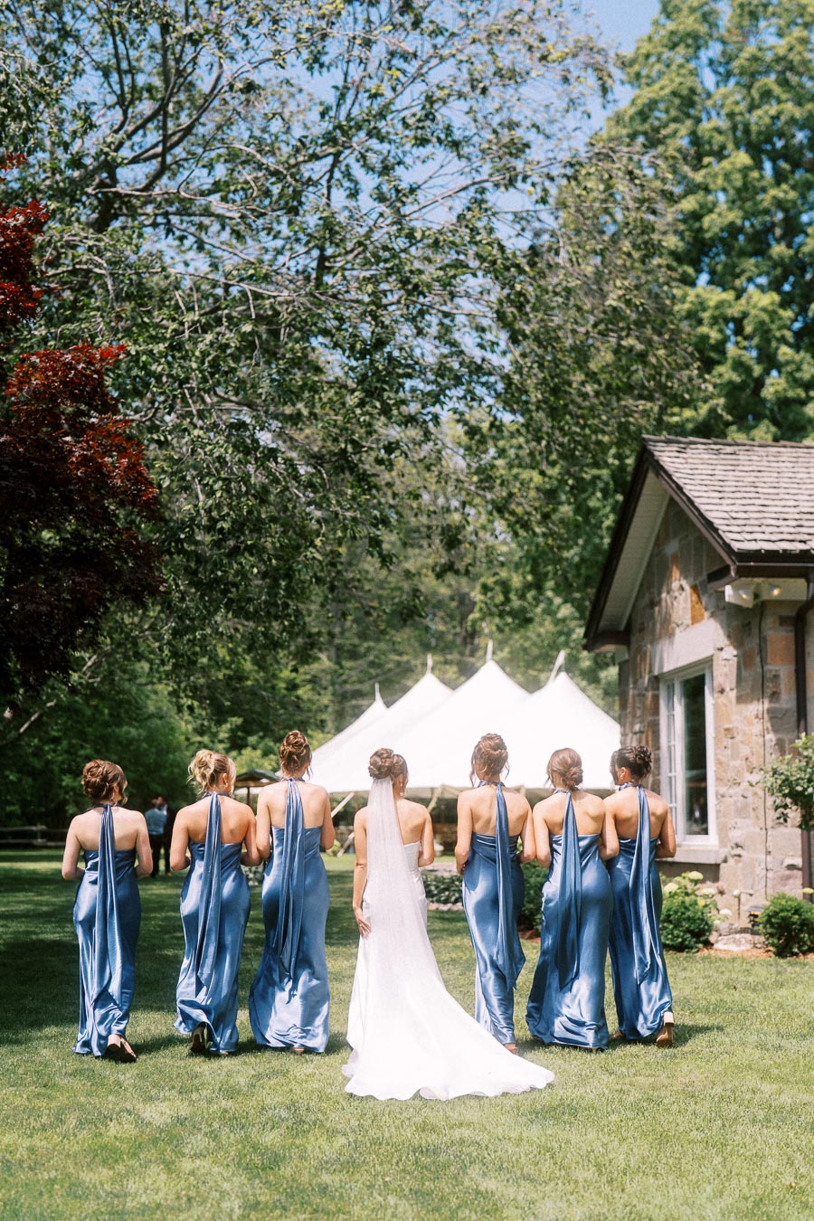Bride with bridesmaids in blue dresses walking across a private estate lawn toward a white wedding tent near Chatham-Kent, Ontario.