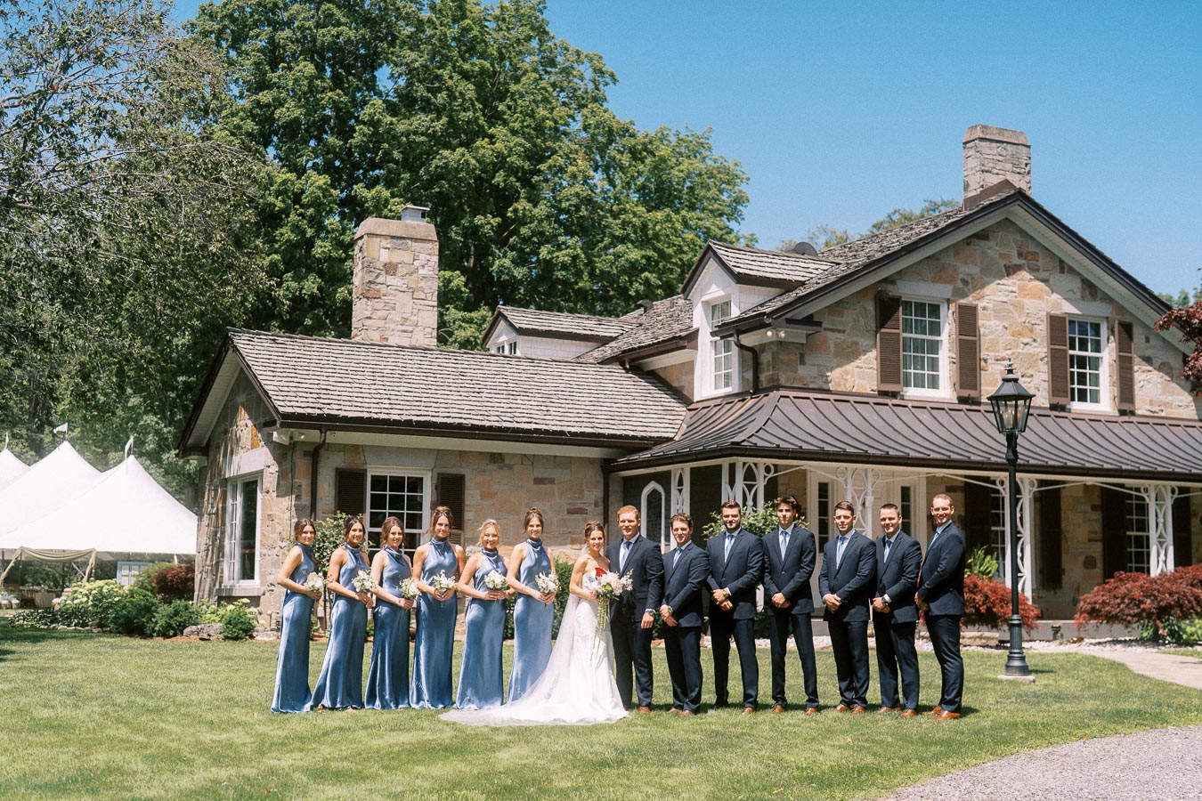 Bride, groom, bridesmaids and groomsmen posing on a lawn in front of a stone estate house with a white wedding tent near Chatham-Kent, Ontario.