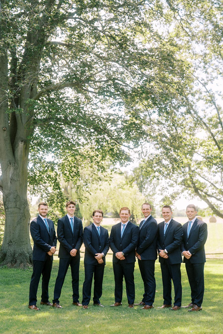 Groomsmen in navy suits standing on grass under a large tree during an outdoor wedding party photo near Chatham-Kent, Ontario.