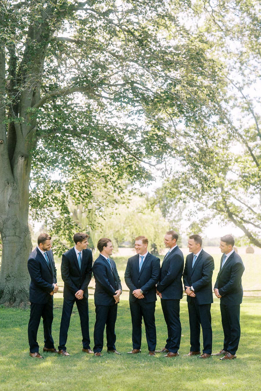 Seven groomsmen in navy suits standing on a lawn under a large tree near Chatham-Kent, Ontario.