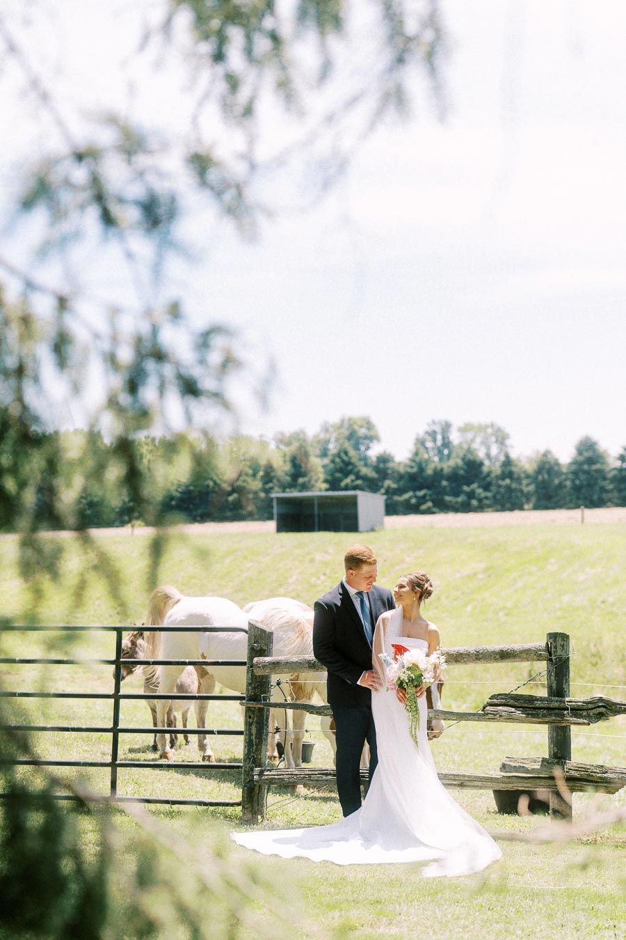 Bride and groom posing by a rustic fence with horses on a private farm property near Chatham-Kent, Ontario, Canada.