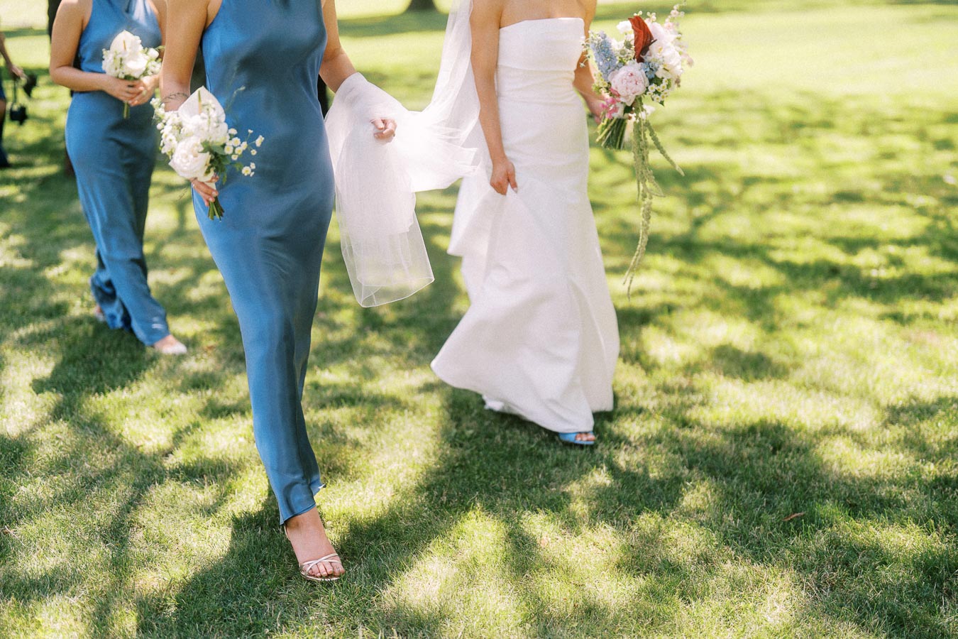 Bride and bridesmaids in blue dresses walking on a sunny lawn with wedding bouquets during an outdoor private estate wedding near Chatham-Kent, Ontario.