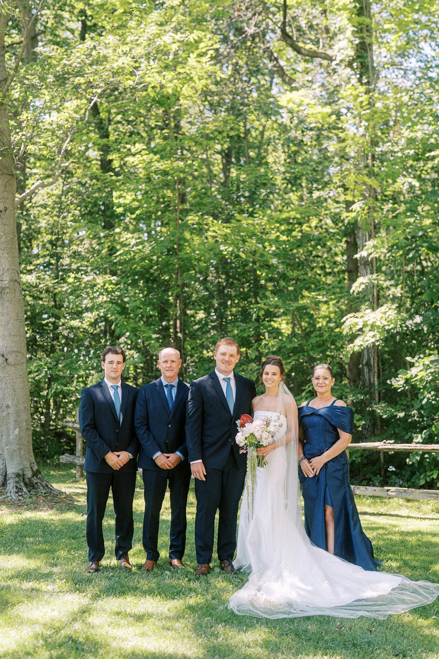 Bride and groom pose with relatives for an outdoor wedding family portrait in a green wooded backyard near Chatham-Kent, Ontario, Canada.
