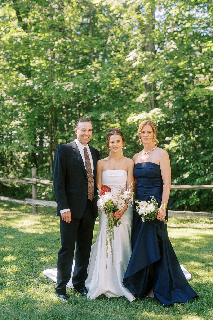 Bride standing with two relatives in formal wedding attire on a grassy private estate near Chatham-Kent, Ontario, with trees and a rustic fence behind them.