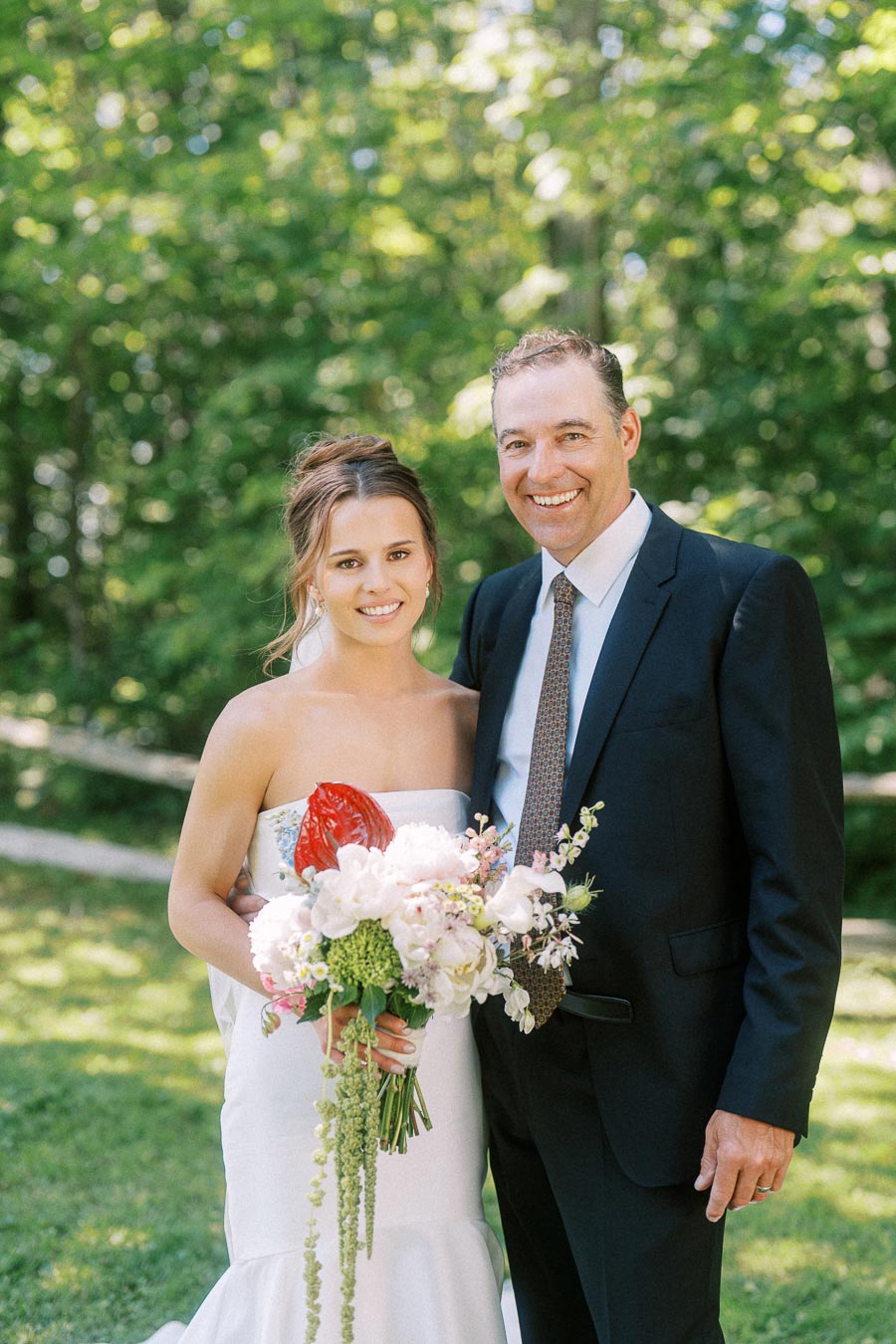 Bride in a white strapless wedding dress holding a floral bouquet beside a smiling man in a black suit outdoors near Chatham-Kent, Ontario.