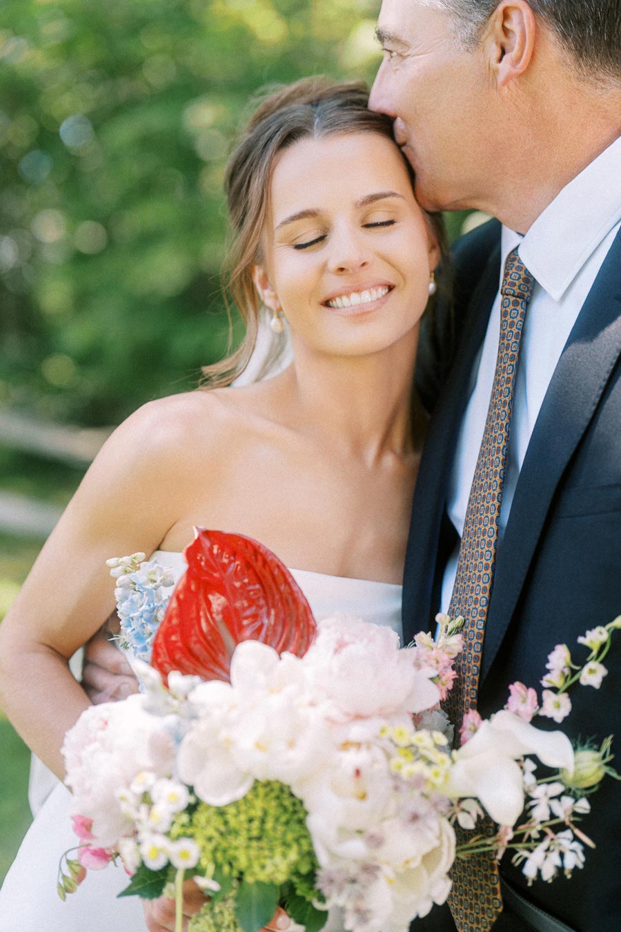 Bride smiles with eyes closed while an older man kisses her forehead during an outdoor wedding portrait near Chatham-Kent, Ontario.