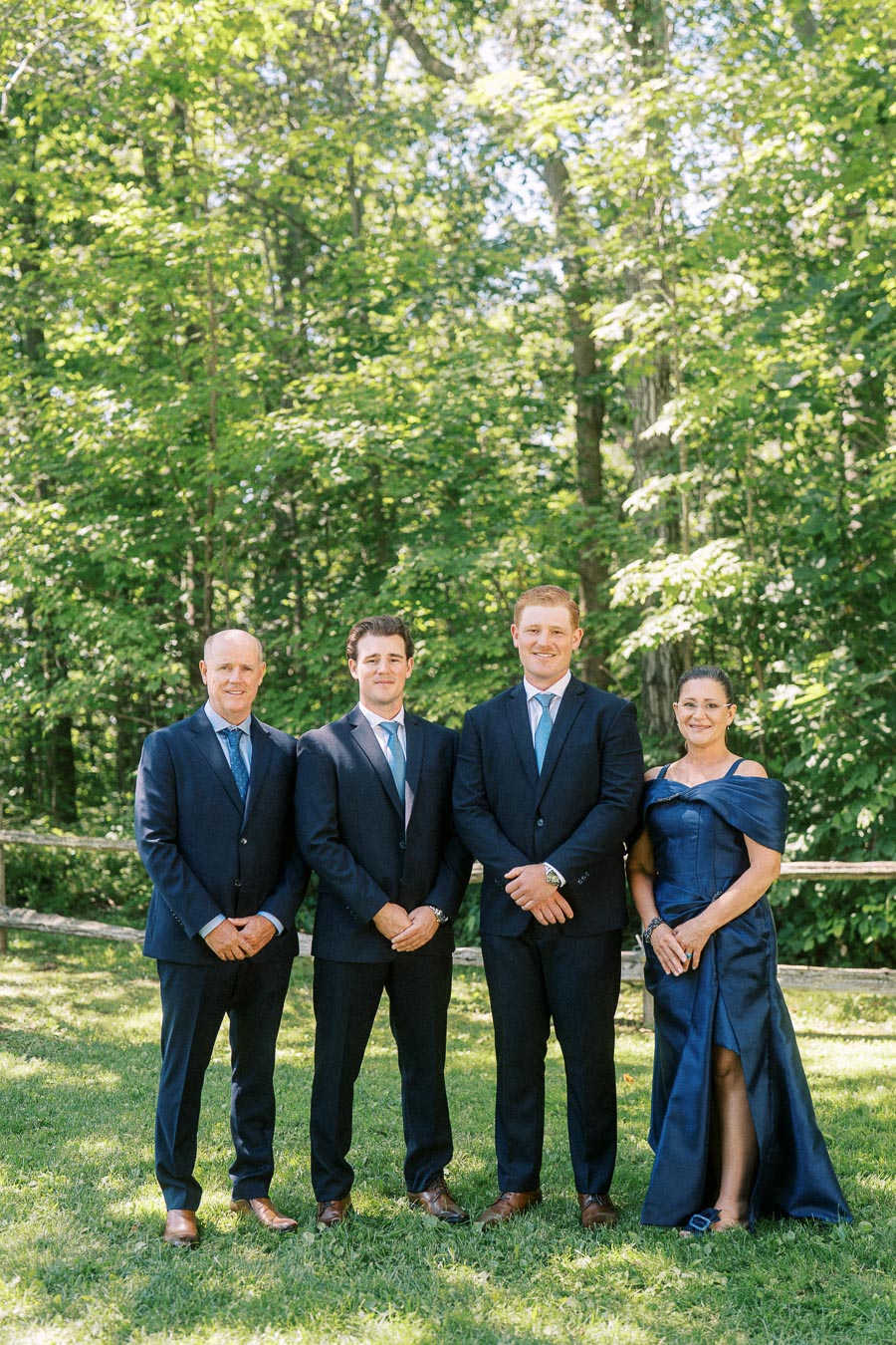 Four wedding guests pose outdoors on grass beside a wooden fence and leafy trees near Chatham-Kent, Ontario, Canada.