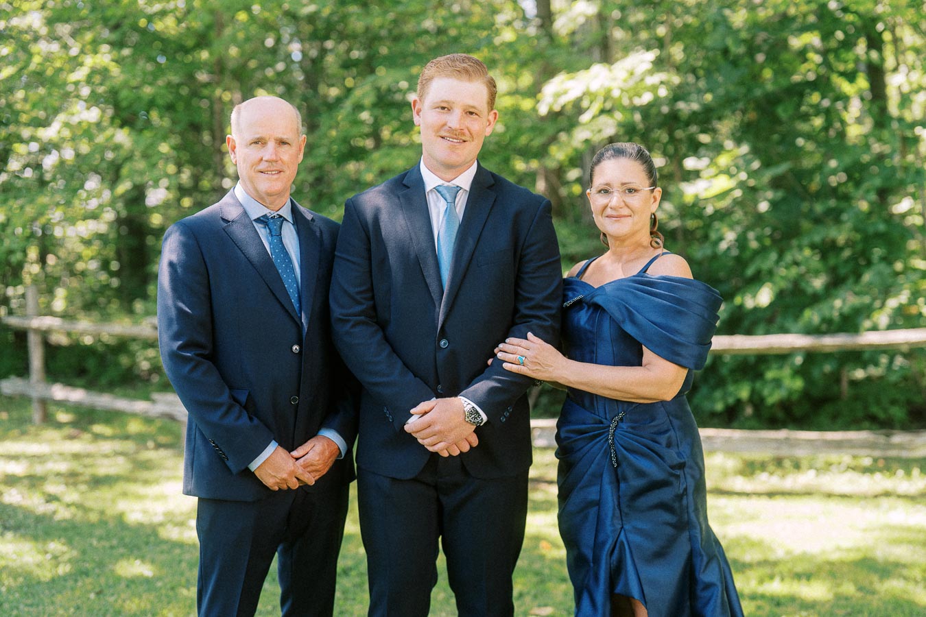 Three wedding guests in navy formal attire posing outdoors near Chatham-Kent, Ontario, with trees, grass, and a rustic wooden fence.