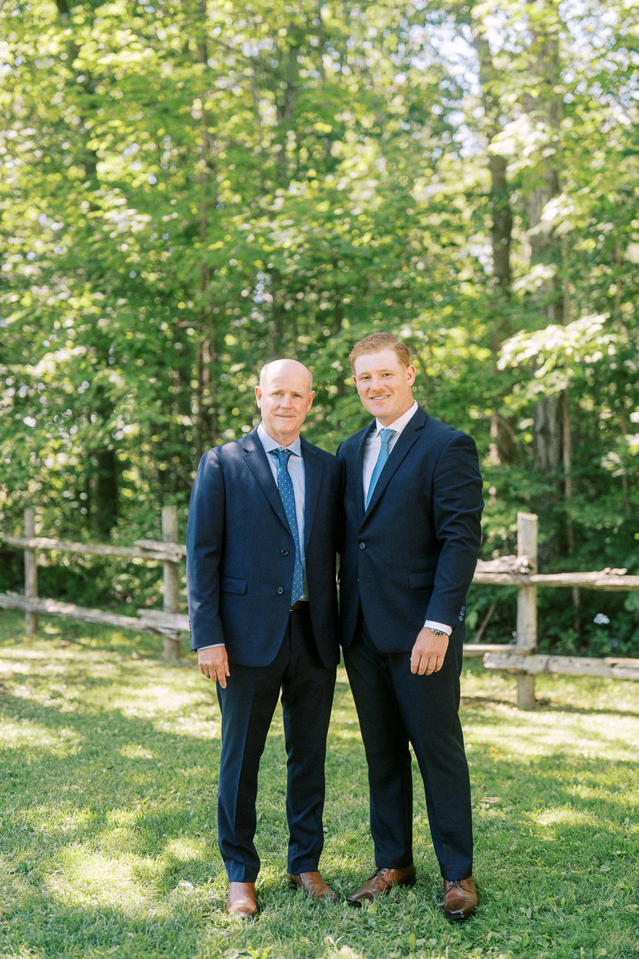 Two men in navy suits pose outdoors on grass near a rustic wooden fence and green trees at a private estate wedding in Chatham-Kent, Ontario.