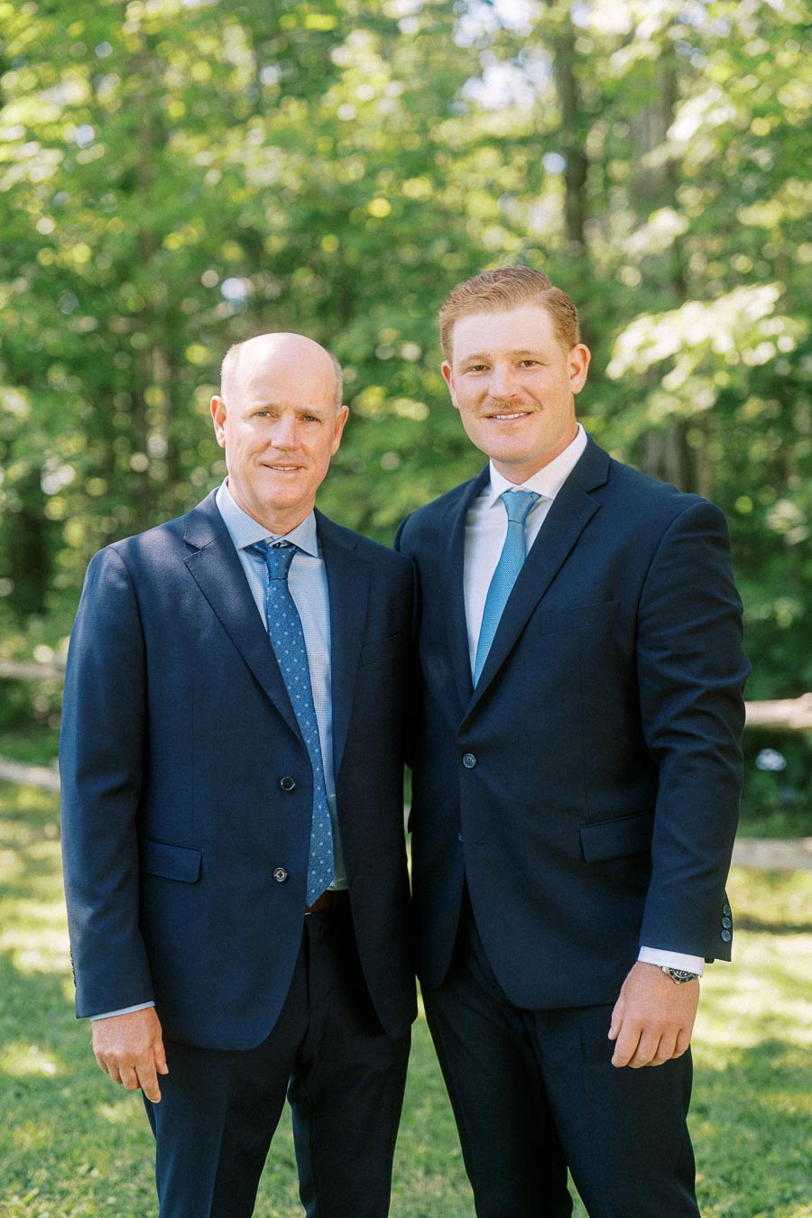 Two men in navy suits pose together outdoors on a grassy lawn with green trees in Chatham-Kent, Ontario.