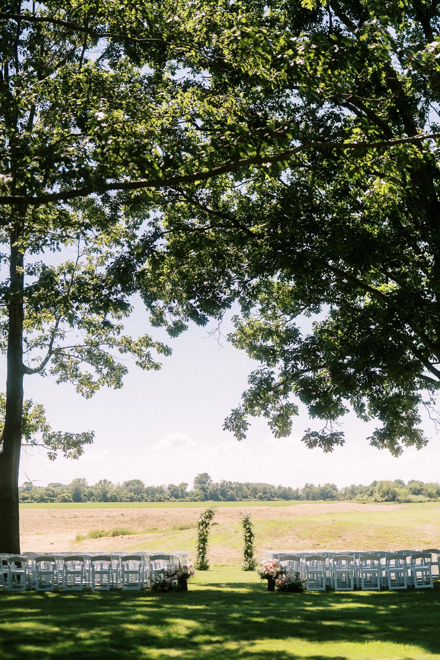 Outdoor private estate wedding ceremony setup with white chairs, floral arch, large shade trees, and farm field near Chatham-Kent, Ontario, Canada.