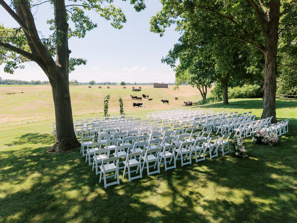 Outdoor wedding ceremony seating with white chairs and floral arbor on a shaded lawn beside a farm field near Chatham-Kent, Ontario.