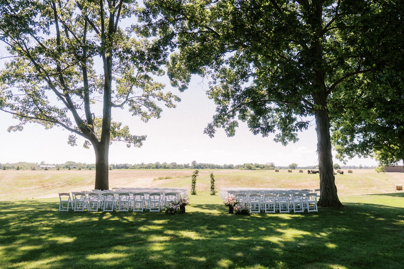 Outdoor wedding ceremony setup with white chairs and floral aisle under large trees overlooking farmland near Chatham Kent, Ontario, Canada.