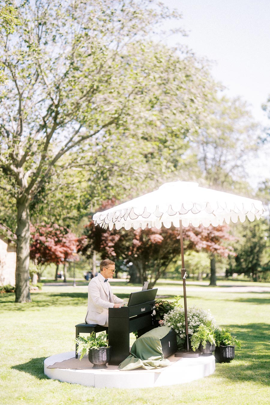 Pianist in a white jacket playing a black Roland digital piano under a white parasol on a green lawn at an outdoor estate wedding in Ontario.