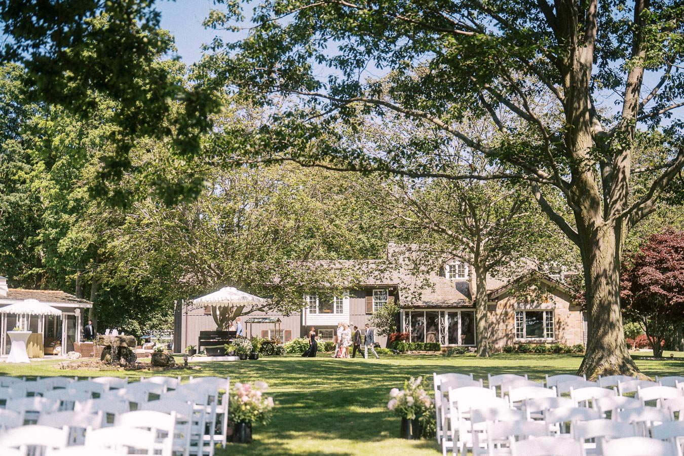 Outdoor private estate wedding setup with white chairs, cocktail hour guests, tent, farmhouse, and large trees near Chatham-Kent, Ontario.