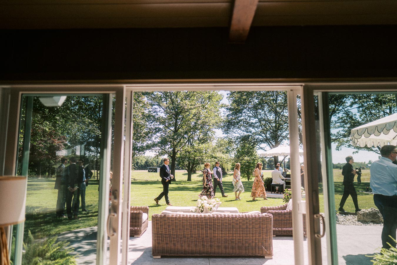 Wedding guests walk across a green lawn outside a private estate near Chatham-Kent, Ontario, viewed through open patio doors during cocktail hour.