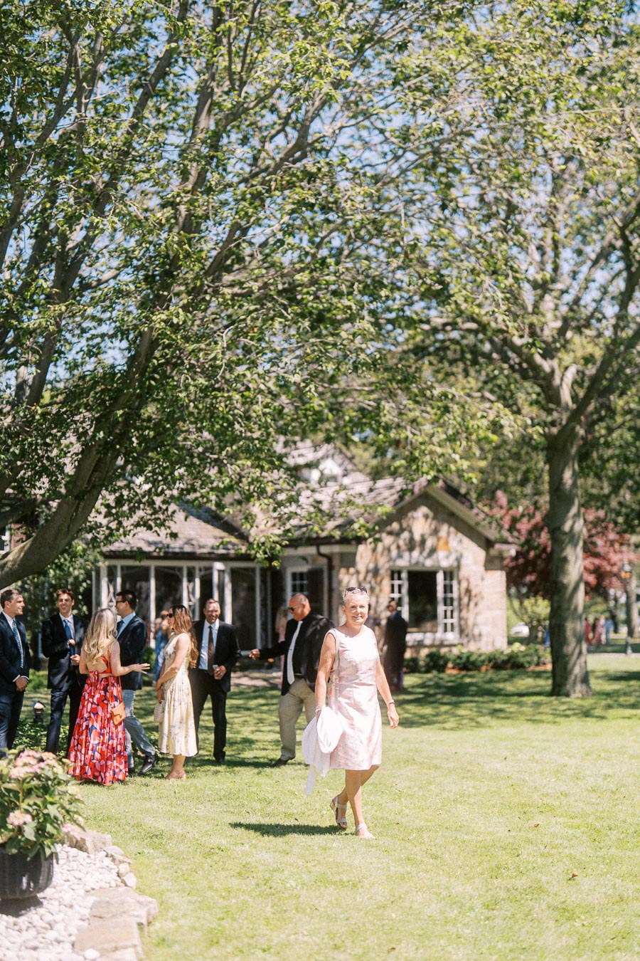 Wedding guests mingle on a sunny lawn during cocktail hour at a private estate near Chatham-Kent, Ontario, with trees and a house in the background.