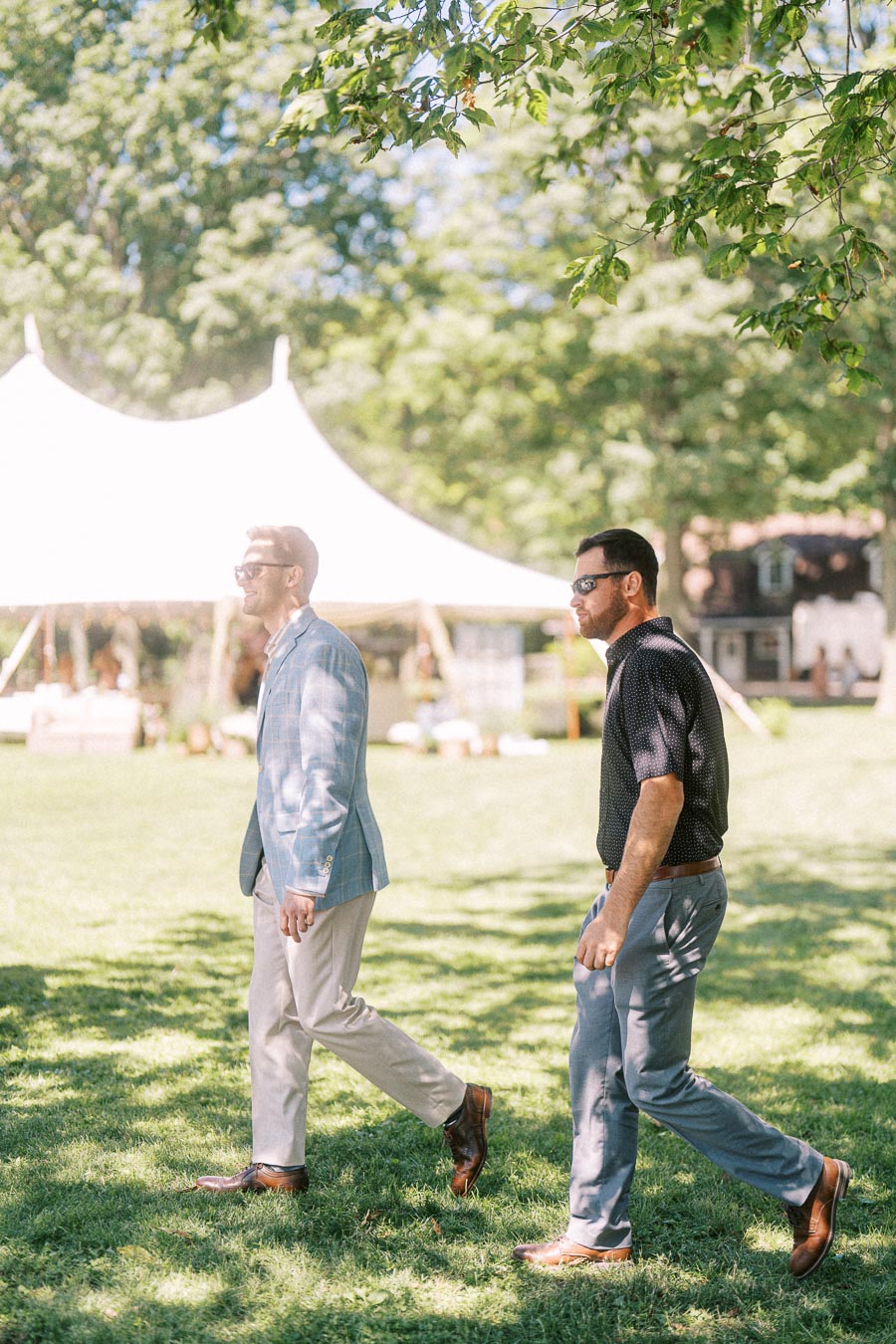 Two male wedding guests in sunglasses walking across a sunny lawn near a white event tent at a private estate wedding in Chatham-Kent, Ontario.
