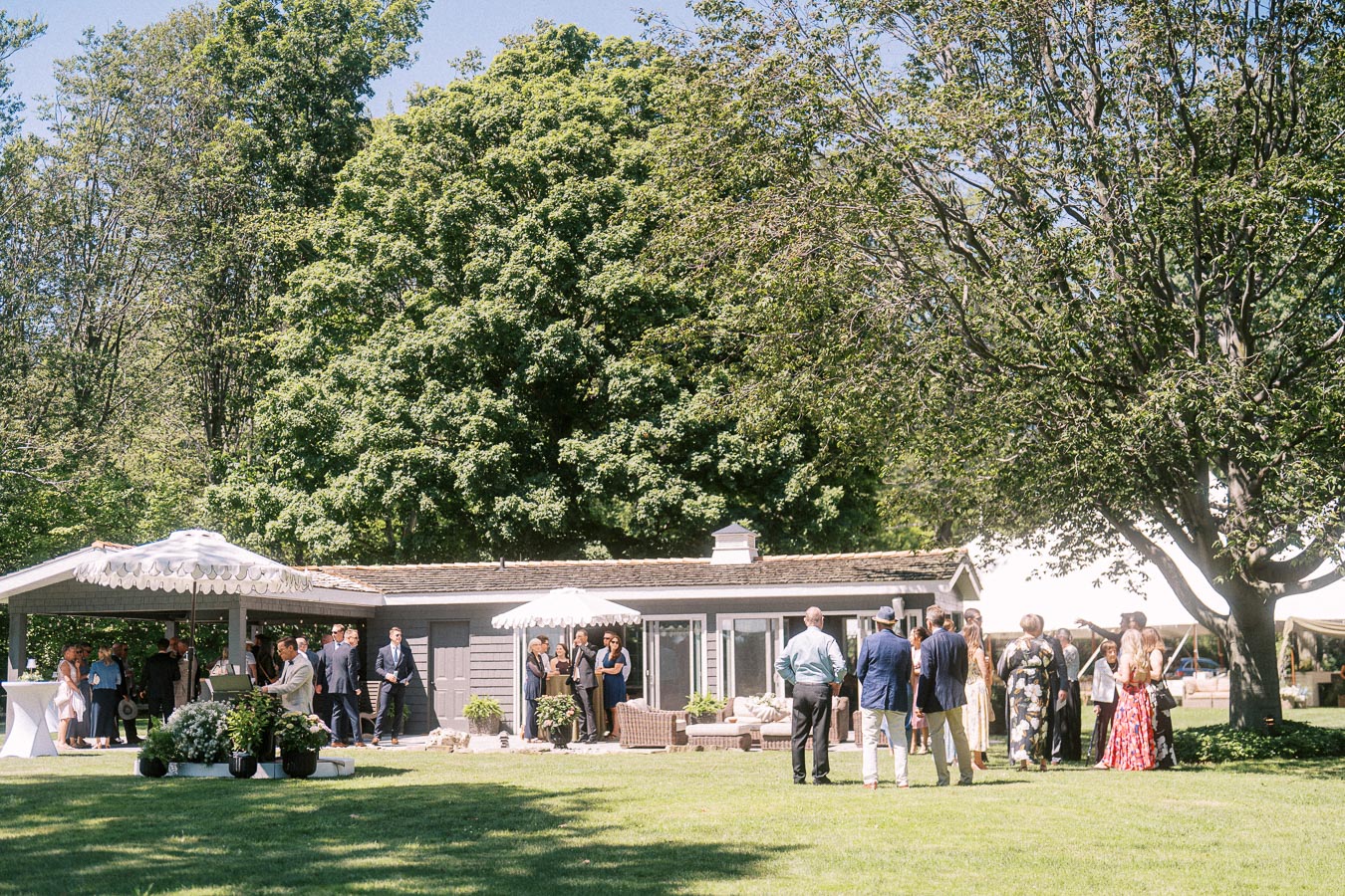 Wedding guests gather for cocktail hour on a lawn at a private estate near Chatham-Kent, Ontario, with umbrellas, patio seating, and trees.