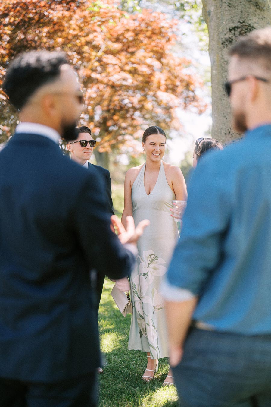 Wedding guests talking during cocktail hour outdoors on a private estate near Chatham-Kent, Ontario, with trees, grass, and summer sunlight.