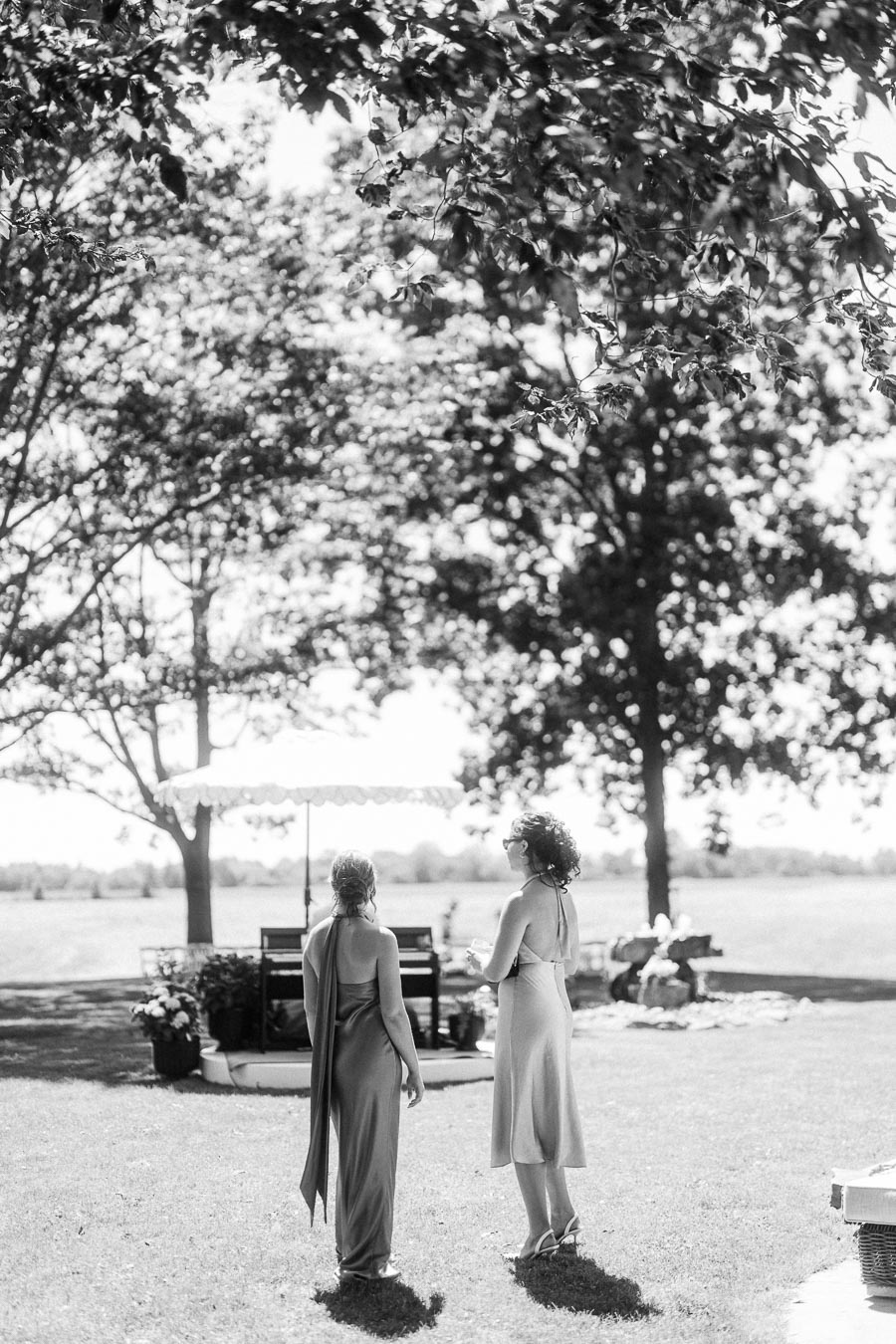 Black and white photo of two wedding guests standing on a lawn under large trees during an outdoor cocktail hour near Chatham-Kent, Ontario.