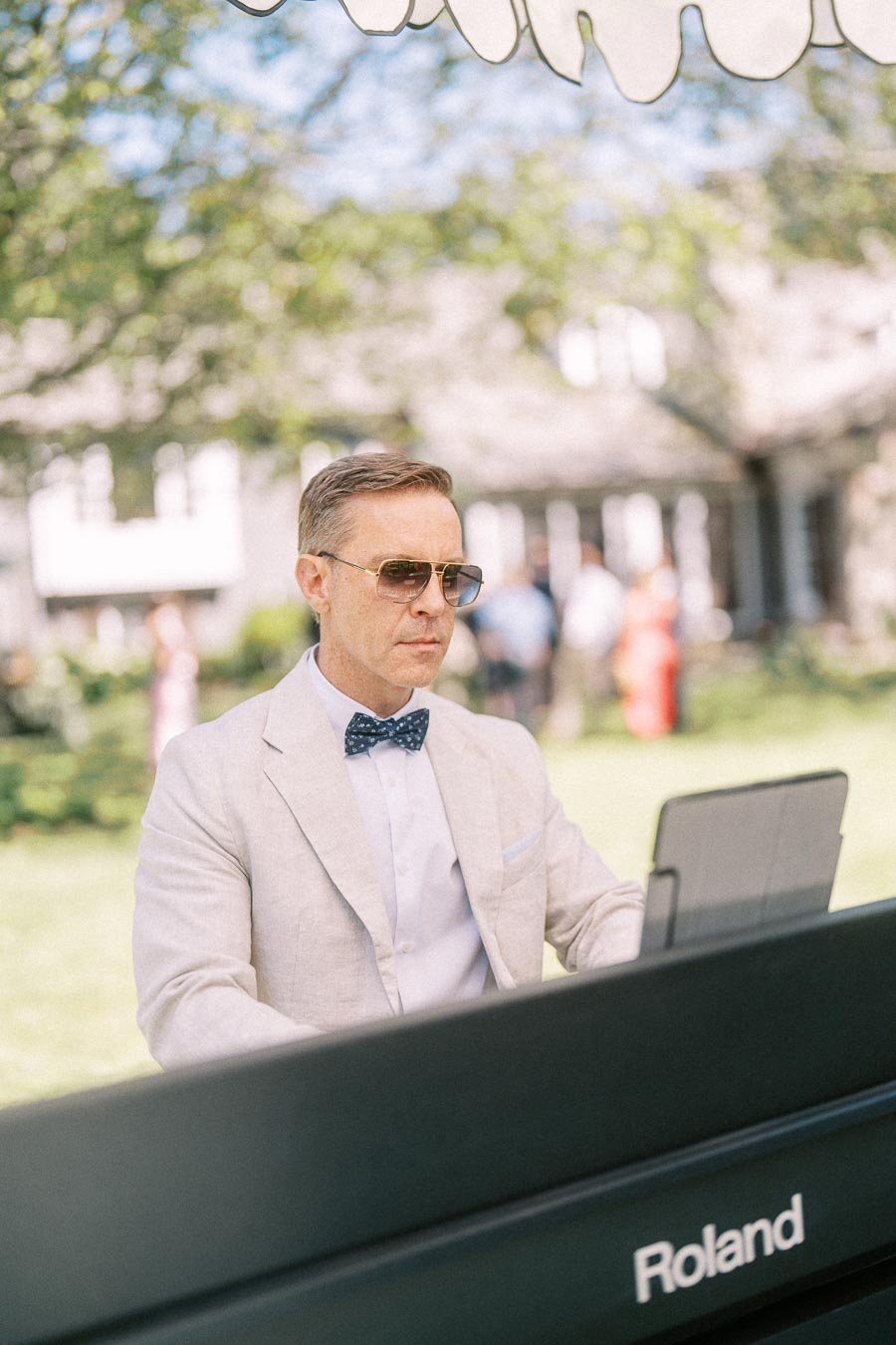 Man in a light suit and sunglasses plays a Roland keyboard outdoors during a private estate wedding cocktail hour near Chatham-Kent, Ontario.