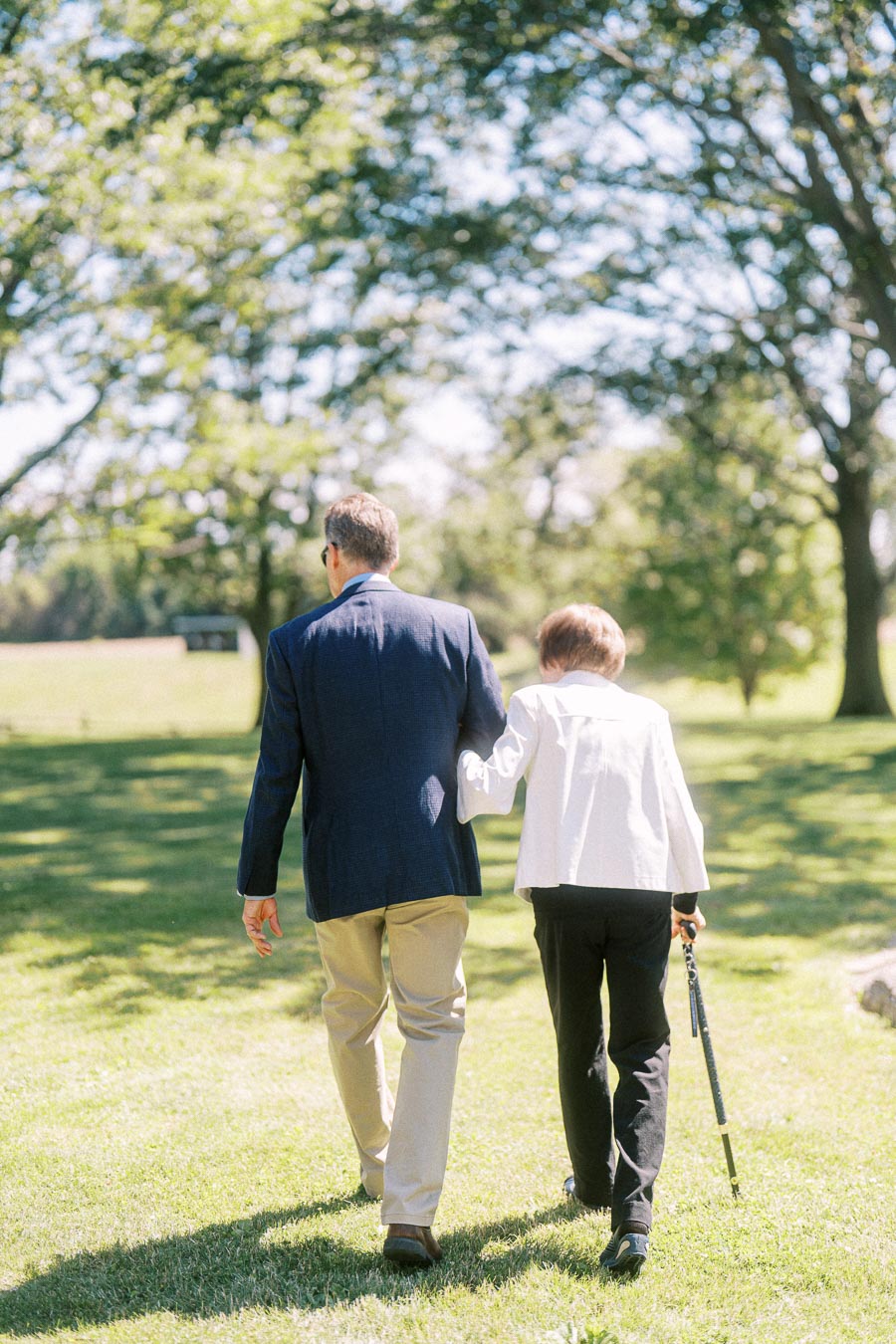 Two wedding guests walk together across a sunny lawn on a private estate near Chatham-Kent, Ontario, with trees and summer greenery.